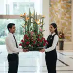 Dos empleados de hotel con uniforme formal sostienen un gran arreglo floral con rosas rojas y aves del paraíso en el lobby luminoso de un edificio