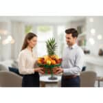 Mujer y hombre jóvenes sosteniendo juntos una canasta de mimbre llena de frutas variadas, con una piña al centro, en una sala de estar moderna y desenfocada