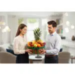 Mujer y hombre jóvenes sosteniendo juntos una canasta de mimbre llena de frutas variadas, con una piña al centro, en una sala de estar moderna y desenfocada