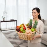 Mujer joven con traje beige sosteniendo una gran canasta de mimbre llena de frutas variadas como piñas, uvas, manzanas y naranjas en una sala moderna y luminosa