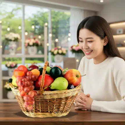 Mujer joven sentada en una mesa de madera mirando una canasta de mimbre llena de frutas variadas como uvas, manzanas, peras y cítricos en una sala luminosa con ventanas y flores de fondo
