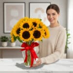 Mujer joven sonriendo junto a un ramo de girasoles amarillos en un florero de cristal con lazo rojo sobre mesa de mármol