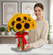 Mujer joven sonriendo junto a un ramo de girasoles amarillos en un florero de cristal con lazo rojo sobre mesa de mármol