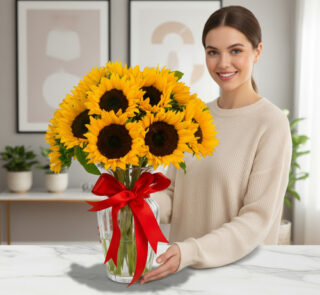Mujer joven sonriendo junto a un ramo de girasoles amarillos en un florero de cristal con lazo rojo sobre mesa de mármol