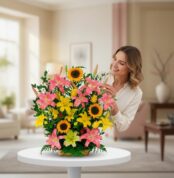 Mujer sonriendo mientras acomoda un arreglo floral con girasoles y lirios rosados y amarillos sobre una mesa blanca en una sala de estar luminosa