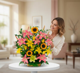 Mujer sonriendo mientras acomoda un arreglo floral con girasoles y lirios rosados y amarillos sobre una mesa blanca en una sala de estar luminosa