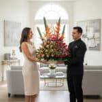 Mujer y repartidor sostienen un gran arreglo floral con rosas y aves del paraíso en una sala de estar luminosa y moderna