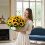 Mujer joven con vestido blanco sosteniendo una canasta de mimbre llena de girasoles amarillos en un salón elegante con sofá azul y grandes ventanales al fondo