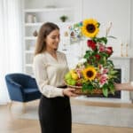 Mujer joven sonriendo mientras recibe una canasta de mimbre con girasoles, rosas rojas, lirios y frutas variadas, decorada con un globo, en una sala de estar luminosa