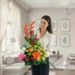 Mujer joven de pie en una sala luminosa sosteniendo un arreglo de flores tropicales naranjas y rojas combinado con frutas variadas y un peluche blanco sobre una base verde