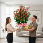 Hombre entrega a mujer un gran arreglo de rosas rojas con lirios rosas, globo de te amo y caja de chocolates en una sala de estar luminosa