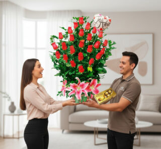 Hombre entrega a mujer un gran arreglo de rosas rojas con lirios rosas, globo de te amo y caja de chocolates en una sala de estar luminosa