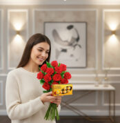 Mujer sonriente con suéter beige sosteniendo un ramo de rosas rojas y una caja de bombones dorados en una sala elegante desenfocada