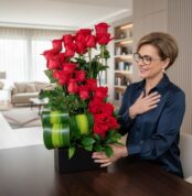 Mujer de mediana edad con blusa azul y gafas sonriendo mientras observa un arreglo de rosas rojas en una base negra sobre una mesa, en una sala de estar luminosa y moderna