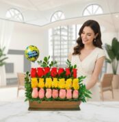 Mujer sonriente junto a un arreglo de rosas rojas, amarillas y rosas en base de madera con globo de Happy Birthday en una sala luminosa