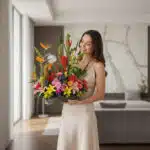 mujer joven con vestido beige sosteniendo un gran arreglo floral tropical de colores vivos en el interior de una sala moderna y luminosa