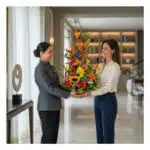 Mujer con uniforme gris entrega a otra mujer un arreglo floral alto y colorido en una sala moderna y luminosa