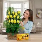 Mujer joven sonriendo mientras acomoda un arreglo de rosas amarillas en base de tronco junto a una caja de chocolates Ferrero Rocher sobre una mesa en una sala moderna