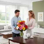 Hombre y mujer de pie junto a una mesa de madera observando un gran ramo de gerberas de colores en un jarrón de cristal en un salón luminoso