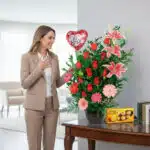 Mujer de traje beige sonríe frente a un gran arreglo floral con rosas rojas, lirios rosados, gerberas y globo de corazón que dice te amo junto a caja de chocolates sobre una mesa