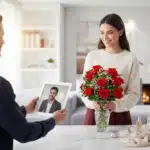 Mujer joven sonríe junto a un ramo de rosas rojas en un jarrón de cristal mientras una persona le muestra una videollamada en una tablet en una sala moderna