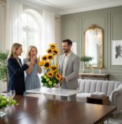 Hombre con traje claro entregando un arreglo alto de girasoles a dos mujeres en un salón moderno y luminoso