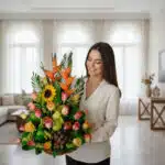 Mujer joven de camisa clara sosteniendo un gran arreglo de flores y frutas con girasoles, rosas de colores, aves del paraíso, uvas y peras en una sala de estar luminosa