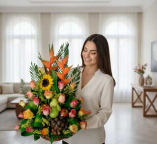 Mujer joven de camisa clara sosteniendo un gran arreglo de flores y frutas con girasoles, rosas de colores, aves del paraíso, uvas y peras en una sala de estar luminosa