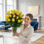 Mujer joven sonriente con camisa beige sosteniendo un gran ramo de rosas amarillas en un jarrón de cristal dentro de un salón moderno con sofás azules y chimenea al fondo