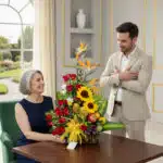 Mujer mayor de cabello canoso y hombre joven de traje beige junto a un arreglo floral con girasoles, rosas rojas, ave del paraíso y frutas sobre una mesa de madera en una sala elegante