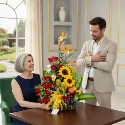 Mujer mayor de cabello canoso y hombre joven de traje beige junto a un arreglo floral con girasoles, rosas rojas, ave del paraíso y frutas sobre una mesa de madera en una sala elegante