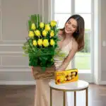 Mujer joven con vestido beige sosteniendo un arreglo de rosas amarillas en base de madera junto a una caja de chocolates Ferrero Rocher sobre una mesa redonda blanca en una sala luminosa