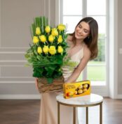 Mujer joven con vestido beige sosteniendo un arreglo de rosas amarillas en base de madera junto a una caja de chocolates Ferrero Rocher sobre una mesa redonda blanca en una sala luminosa