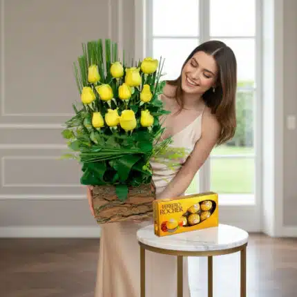 Mujer joven con vestido beige sosteniendo un arreglo de rosas amarillas en base de madera junto a una caja de chocolates Ferrero Rocher sobre una mesa redonda blanca en una sala luminosa