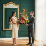 Hombre con traje oscuro y mujer con vestido crema sostienen juntos un arreglo floral tropical de colores vivos en una sala de paredes verde oscuro con gran espejo dorado