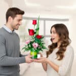 Hombre joven con suéter gris entrega un arreglo floral con rosas rojas y margaritas rosadas a una mujer sorprendida de cabello largo castaño en una sala luminosa