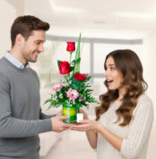 Hombre joven con suéter gris entrega un arreglo floral con rosas rojas y margaritas rosadas a una mujer sorprendida de cabello largo castaño en una sala luminosa