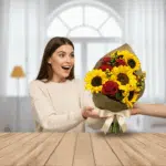 Mujer joven con suéter beige recibe sonriente un ramo de flores con girasoles y rosas rojas sobre una mesa de madera en interior luminoso
