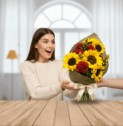 Mujer joven con suéter beige recibe sonriente un ramo de flores con girasoles y rosas rojas sobre una mesa de madera en interior luminoso