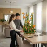 pareja joven de pie junto a una mesa de comedor moderna observando un gran arreglo floral con aves del paraíso y rosas naranjas