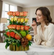 Mujer joven sonriente observa un arreglo floral con rosas naranjas y amarillas combinado con manzana, naranja y uvas sobre una mesa de mármol en sala moderna
