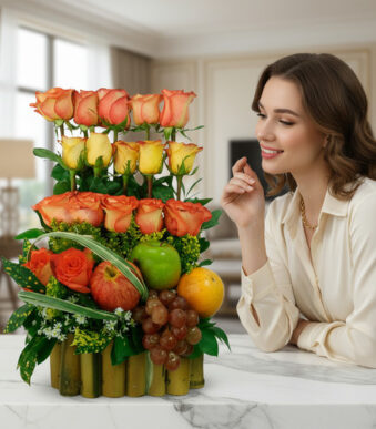 Mujer joven sonriente observa un arreglo floral con rosas naranjas y amarillas combinado con manzana, naranja y uvas sobre una mesa de mármol en sala moderna