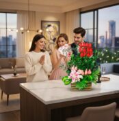 Hombre y mujer entregan a una amiga un arreglo floral con rosas rojas, globos y lirios sobre una isla de cocina en un departamento moderno con vista a la ciudad