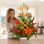 Mujer sonriente sosteniendo un gran arreglo floral tropical con girasoles, lirios naranjas, aves del paraíso y hojas verdes sobre una mesa en una sala luminosa