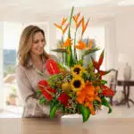 Mujer sonriente sosteniendo un gran arreglo floral tropical con girasoles, lirios naranjas, aves del paraíso y hojas verdes sobre una mesa en una sala luminosa
