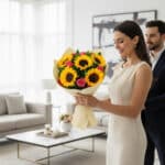 Mujer elegante con vestido beige sosteniendo un ramo de girasoles y rosas en una sala de estar luminosa, con un hombre de traje oscuro detrás de ella