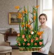 Mujer joven sosteniendo un gran arreglo floral con rosas naranjas y flores aves del paraíso en una sala elegante