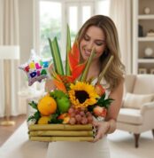 Mujer sonriente sostiene un arreglo de frutas y flores tropicales con girasol y globo que dice feliz día en una sala de estar luminosa