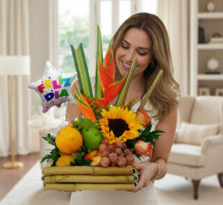 Mujer sonriente sostiene un arreglo de frutas y flores tropicales con girasol y globo que dice feliz día en una sala de estar luminosa