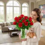 Mujer joven sostiene un jarrón de cristal con un ramo de rosas rojas y un oso de peluche marrón claro en una sala de estar luminosa con sofá y grandes ventanales al fondo